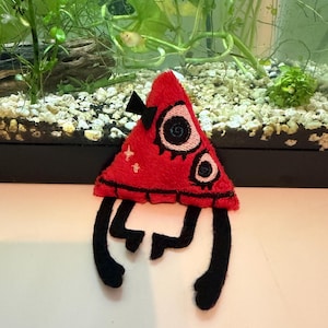 Könnte beinhalten: Ein rotes, dreieckiges Plüschtier mit schwarzen Details, einschließlich Augen, einer Schleife und Beinen. Das Spielzeug hat ein skurriles Design und befindet sich vor einem Aquarium mit Pflanzen und kleinen Fischen.