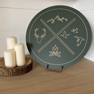 May include: A dark green round wooden sign with a white background and gold accents. The sign features a deer, mountains, a campfire, and two birds. The sign is on a wooden stand.