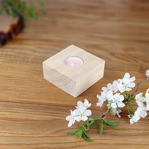 May include: A light wood candle holder with a square base and a round opening for a tea light. A pink tea light is in the holder. A branch of white blossoms is on a wooden surface next to the holder.