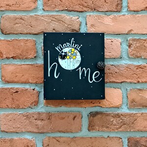 Può includere: Un cartello nero in ardesia con lettere bianche che recita "marlini home" con un gatto nero e fiori gialli in un cerchio sul cartello. Il cartello è appeso a un muro di mattoni.
