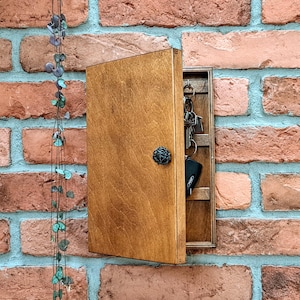 Op de afbeelding: Een houten sleutelhouder met een scharnierende deur, gemonteerd op een bakstenen muur. De sleutelhouder is open en onthult sleutels die erin hangen. De deur heeft een decoratieve zwarte knop. Het hout heeft een warme, natuurlijke afwerking.