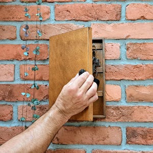 May include: A wooden key holder mounted on a brick wall. The key holder is open, revealing keys inside. The wood has a natural finish. A hand is opening the key holder. A plant with small, round leaves hangs on the left.