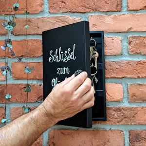 Pode incluir: Um porta-chaves preto montado numa parede de tijolo. O porta-chaves tem as palavras "Schlüssel zum Glück" em escrita branca. Várias chaves penduram-se dentro do porta-chaves aberto. Uma mão está a abrir o porta-chaves.