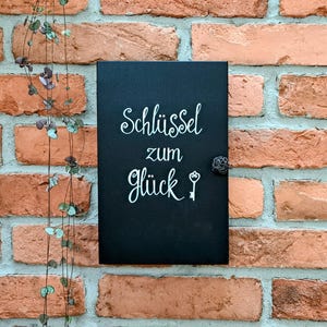 May include: A black rectangular sign with the words "Schl&uuml;ssel zum Gl&uuml;ck" in white script, featuring a key illustration. The sign has a small, decorative knob and is mounted on a red brick wall. A plant with small green leaves hangs on the left.