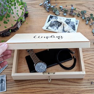 May include: A wooden box with a lid that says "Unplug" on the top. The box is open and contains a watch, a phone, and a bracelet.