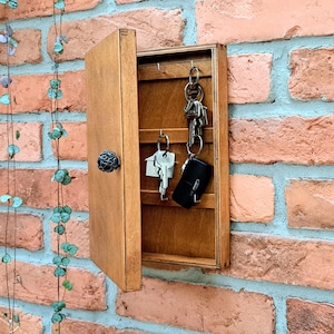 May include: A wooden key holder with a hinged door, mounted on a brick wall. The key holder is open, revealing keys hanging inside. The door has a decorative black knob. A string of decorative leaves hangs on the left.