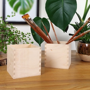 Peut inclure: Deux porte-crayons carrés en bois naturel avec un ensemble de pinceaux en arrière-plan. Les porte-crayons sont en bois clair et ont une finition naturelle.