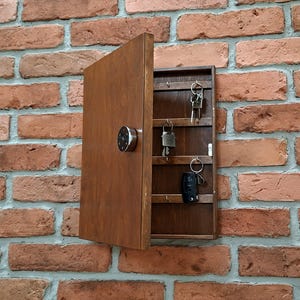 May include: A rectangular wooden key cabinet with a hinged door and a circular silver knob. The interior features three horizontal shelves with metal hooks, currently holding several keys and a black key fob, mounted on a textured brick wall.
