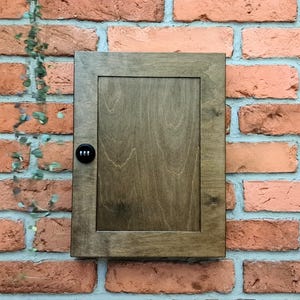 Puede incluir: Una caja de madera marrón oscuro montada en la pared con una cerradura de combinación. La caja está montada en una pared de ladrillo.