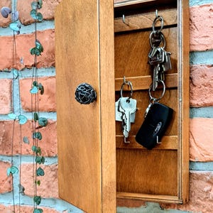 May include: A wooden key holder with a hinged door and a decorative metal knob. The interior has hooks for hanging keys. The key holder is mounted on a red brick wall. Several keys are hanging from the hooks.