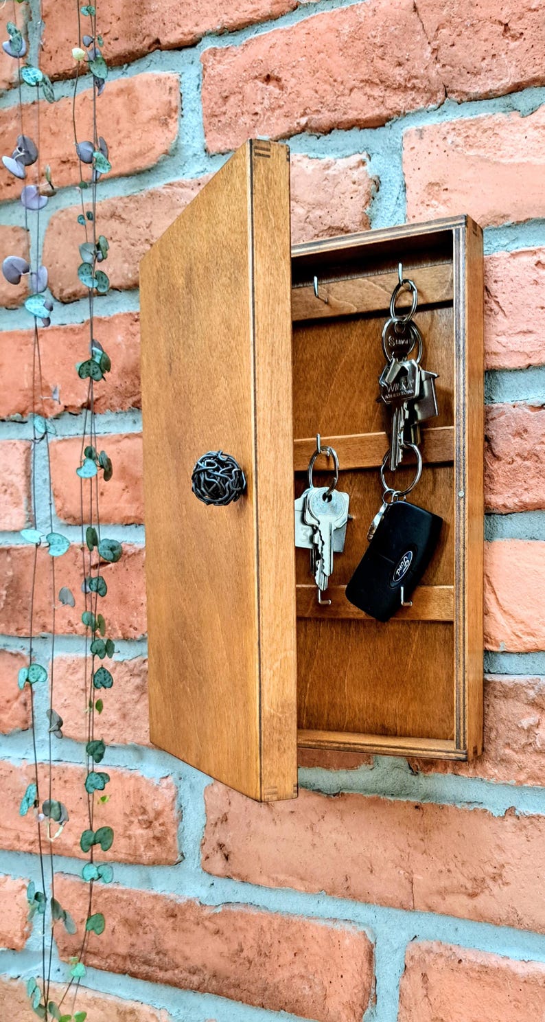 May include: A wooden key holder with a hinged door and a decorative metal knob. The interior has hooks for hanging keys. The key holder is mounted on a red brick wall. Several keys are hanging from the hooks.