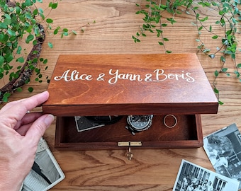 May include: A wooden box with a hinged lid, personalised with the names "Alice & Yann & Boris" in white lettering. The box is open, revealing a watch, a ring, and old photographs inside.
