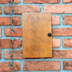 May include: A rectangular wooden cabinet door with a dark knob is mounted on a brick wall. The wood has a warm, natural finish. A string of decorative leaves hangs on the left side of the cabinet.