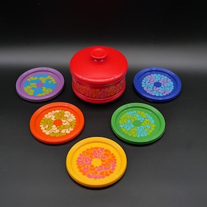 Peut inclure: Un récipient rouge avec couvercle et un ensemble de dessous de verre colorés. Les dessous de verre ronds présentent des motifs floraux violets, bleus, verts, oranges et jaunes. L'ensemble est sur fond noir.