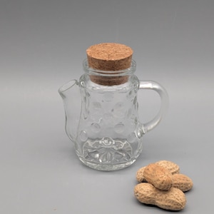 Peut inclure: Un récipient transparent en forme de théière en verre avec un bouchon en liège. Le récipient a une poignée et un bec verseur. Un petit tas de cacahuètes est posé à côté du récipient. Le récipient mesure environ 10 cm de haut.