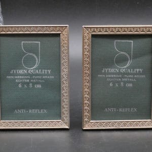Könnte beinhalten: Zwei rechteckige Bilderrahmen aus Messing mit einer geprägten Zierkante. Die dunkelgrünen Einlagen tragen den Text JYDEN QUALITY, REN MESSING - PURE BRASS, ECHTER METALL, 6 x 8 cm und ANTI-REFLEX. Die Rahmen haben eine metallische Oberfläche.