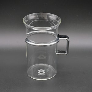 Può includere: Una tazza di vetro trasparente con un manico nero e una fascia metallica al centro. La tazza ha un corpo cilindrico e una parte superiore svasata. Il fondo della tazza riporta la scritta "DENMARK" in un logo circolare.