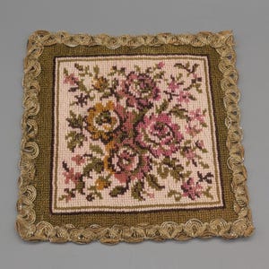 Könnte beinhalten: Quadratisches Nadelspitzentextil mit floralem Design. Cremefarbener Hintergrund mit rosa, gelben und grünen Blumen. Umrandet von einem dunkelgrünen Rand und einer goldfarbenen, gewellten Bordüre. Dekorative Textilie.