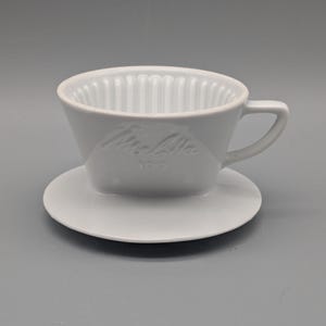 Puede incluir: Un filtro de café de cerámica blanca con un platillo a juego. El filtro tiene estrías verticales en el interior y un asa lateral. La marca "Melitta" está grabada en relieve en la parte delantera, junto con el número "100".
