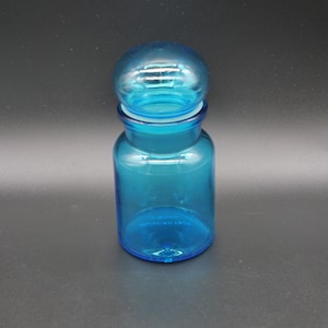 Puede incluir: Un tarro de boticario de vidrio turquesa transparente con tapa redonda. El tarro tiene un cuerpo cilíndrico y una base redondeada. La tapa es de color turquesa a juego y encaja perfectamente. El tarro está vacío.