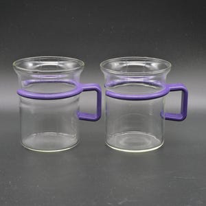 Peut inclure: Deux mugs en verre transparent avec des poignées violettes. Les mugs ont une forme cylindrique et un bord arrondi. Les poignées violettes contrastent avec le verre transparent. Les mugs sont sur fond sombre.