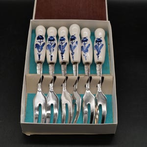 Puede incluir: Un juego de seis tenedores de metal plateado con mangos de cerámica blanca que presentan un patrón floral azul. Los tenedores están dispuestos en una caja rectangular blanca con un forro interior turquesa y una tapa abierta en ángulo.