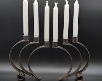 May include: A dark metal candelabra featuring six curved, circular base supports and six vertical candle holders. Each holder contains a slender, white, unscented taper candle. The structure is composed of interconnected metal arcs.
