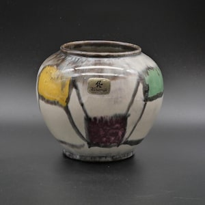 May include: A ceramic vase with a cream-colored base, featuring abstract designs in yellow, green, and purple. The vase has a dark brown rim and a small metal tag that reads "Keramik". The overall shape is round.