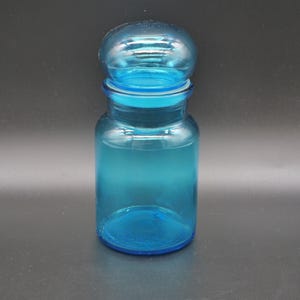 Puede incluir: Un tarro de vidrio turquesa transparente con tapa a juego. El tarro tiene un cuerpo cilíndrico y una tapa redondeada, creando un estilo de boticario clásico. El vidrio es liso y refleja la luz, mostrando el color azul.