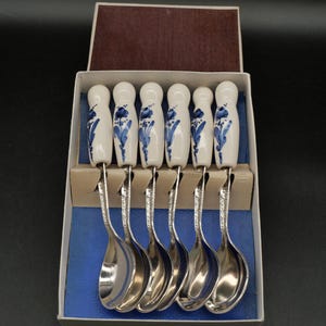 Puede incluir: Un juego de seis cucharas de metal plateado con mangos de cerámica blanca, cada una con un diseño floral azul pintado a mano. Las cucharas están dispuestas una al lado de la otra en una caja de cartón rectangular con una base interior azul.
