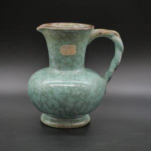 Könnte beinhalten: Türkisfarbener Keramikkrug mit rundem Boden und gebogenem Henkel. Der Krug hat eine strukturierte, gesprenkelte Oberfläche und ein kleines Etikett. Der Rand und der Henkel sind dunkelbraun. Etwa 15 cm hoch.