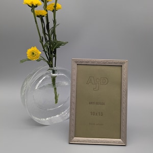 Puede incluir: Un marco de fotos plateado con un borde decorativo se encuentra junto a un jarrón de cristal transparente con flores amarillas. El marco tiene el texto "AJD ANTI REFLEX 10x15". Las flores tienen tallos y hojas verdes. Medidas 10cm x 15cm.