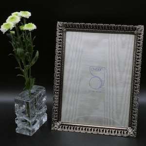 Puede incluir: Un marco de fotos rectangular con un borde de filigrana metálica oscura y ornamentada, y un frente de cristal. Dentro del marco hay un inserto blanco con el texto azul JYDEN. Al lado, un jarrón de cristal transparente y texturizado contiene flores blancas y verdes.