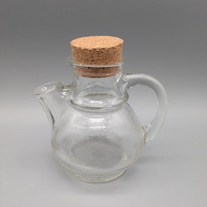 Könnte beinhalten: Ein klares Glas-Ölkännchen mit rundem Körper, kleinem Ausguss und Griff. Das Ölkännchen ist mit einem Korken verschlossen. Das Glas hat eine strukturierte Oberfläche. Das Ölkännchen ist für Flüssigkeiten bestimmt.