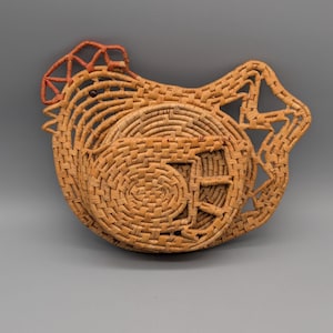 Könnte beinhalten: Ein geflochtener Korb in Form eines Hahns, hergestellt aus naturfarbenem Stroh. Der Korb hat eine kreisförmige Basis mit einem Spiralmuster und einen roten Akzent auf dem Kamm. Der Korb hat ausgeschnittene geometrische Formen.