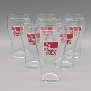 Könnte beinhalten: Sechs klare Gläser mit geschwungener Form. Jedes Glas trägt ein rot-weißes Logo mit der Aufschrift "Sinalco COLA". Die Gläser sind in einem Halbkreis angeordnet, wodurch ihr Design und ihre Klarheit hervorgehoben werden.