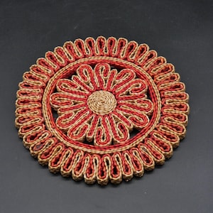 Könnte beinhalten: Runder gewebter Untersetzer mit floralem Design. Der Untersetzer hat ein zentrales Blumenmuster in Rot und Beige, umgeben von einem Ring aus roten und beigen Schlaufen. Hergestellt aus Naturfasern, geeignet für die Inneneinrichtung.