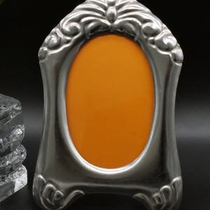 May include: A silver-toned metal picture frame with an arched top and an oval-shaped center opening. The frame features embossed scrollwork and decorative detailing along the top and bottom edges, surrounding a solid orange insert.