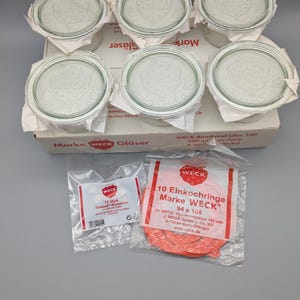 May include: Six clear glass Weck jars with lids, presented in a box. The image also shows two small packets: one with metal clamps and another containing ten orange rubber sealing rings for the jars.