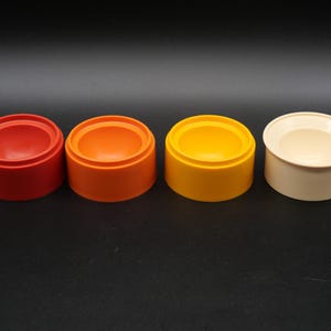 May include: Four colorful, round containers in a row. The containers are red, orange, yellow, and off-white. Each container has a cylindrical base and a recessed, bowl-shaped interior. The containers are set against a black background.