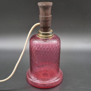 May include: A vintage, small table lamp with a dark brown top and a textured, deep pink glass base. The base has a round, flared bottom and a gold-colored ring. A cream-colored cord extends from the top.