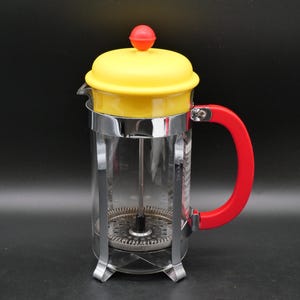 Peut inclure: Une cafetière à piston en verre dotée d'un couvercle jaune vif surmonté d'un petit bouton rouge. La carafe est maintenue par une armature en métal chromé et possède une poignée rouge. Un mécanisme de piston en métal avec filtre est visible.