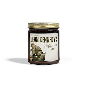 May include: A brown glass jar with a gold lid. The label reads "THIS SMELLS LIKE LEON KENNEDY'S Asserack" with an illustration of a person. The jar is filled with a dark substance.