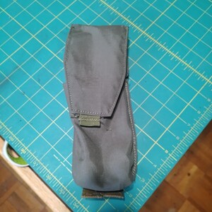 Könnte beinhalten: Eine graue Tasche mit Klappenverschluss und Klettverschluss. Die Tasche besteht aus einem strapazierfähigen Stoff und hat eine rechteckige Form. Der Hintergrund ist eine türkisfarbene Schneidematte mit Rasterlinien.