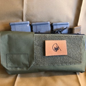 Puede incluir: Una bolsa verde oliva con cremallera en la parte superior, con un parche de cuero con un logotipo. La bolsa contiene varios objetos rectangulares grises. El fondo es una tela beige.