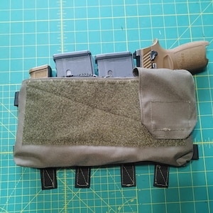 Könnte beinhalten: Eine taktische Brusttasche in Grün- und Beigetönen, zur Aufnahme von Magazinen und einer Handfeuerwaffe. Das Rig verfügt über mehrere Taschen, Klettverschlüsse und verstellbare Riemen. Der Hintergrund ist eine grüne Schneidematte.
