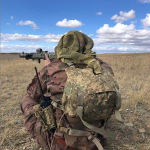 May include: A person in camouflage clothing kneels in a field, aiming a rifle. The person wears a camouflage helmet and a net over their head. A backpack and radio are also visible. The sky is blue with scattered clouds.