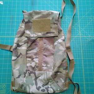 May include: A camouflage backpack with a central zip and a rectangular Velcro patch. The bag has adjustable straps and a flap closure. The colour scheme includes shades of green, brown, and tan, creating a military-inspired aesthetic.