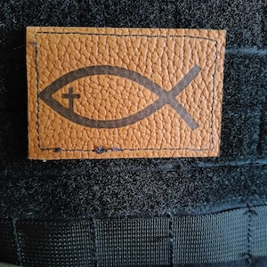 May include: A rectangular, brown leather patch with a stitched border. The patch features a dark brown Ichthys symbol, a Christian symbol of a fish, with a cross inside. The patch is set against a dark, textured background.