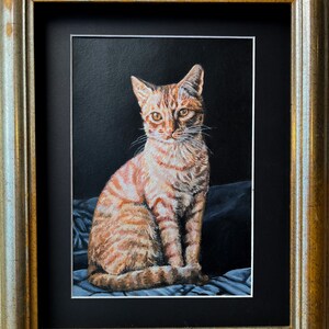 May include: A framed painting of a tabby cat sitting on a blue and white patterned fabric. The cat has orange and white fur and is looking directly at the viewer.